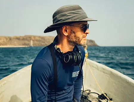 man with hat in boat on the ocean, with headphones around neck and recording equipment